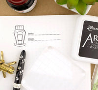 Black and white pen on a white surface with a branded ink pad and decorative elements.