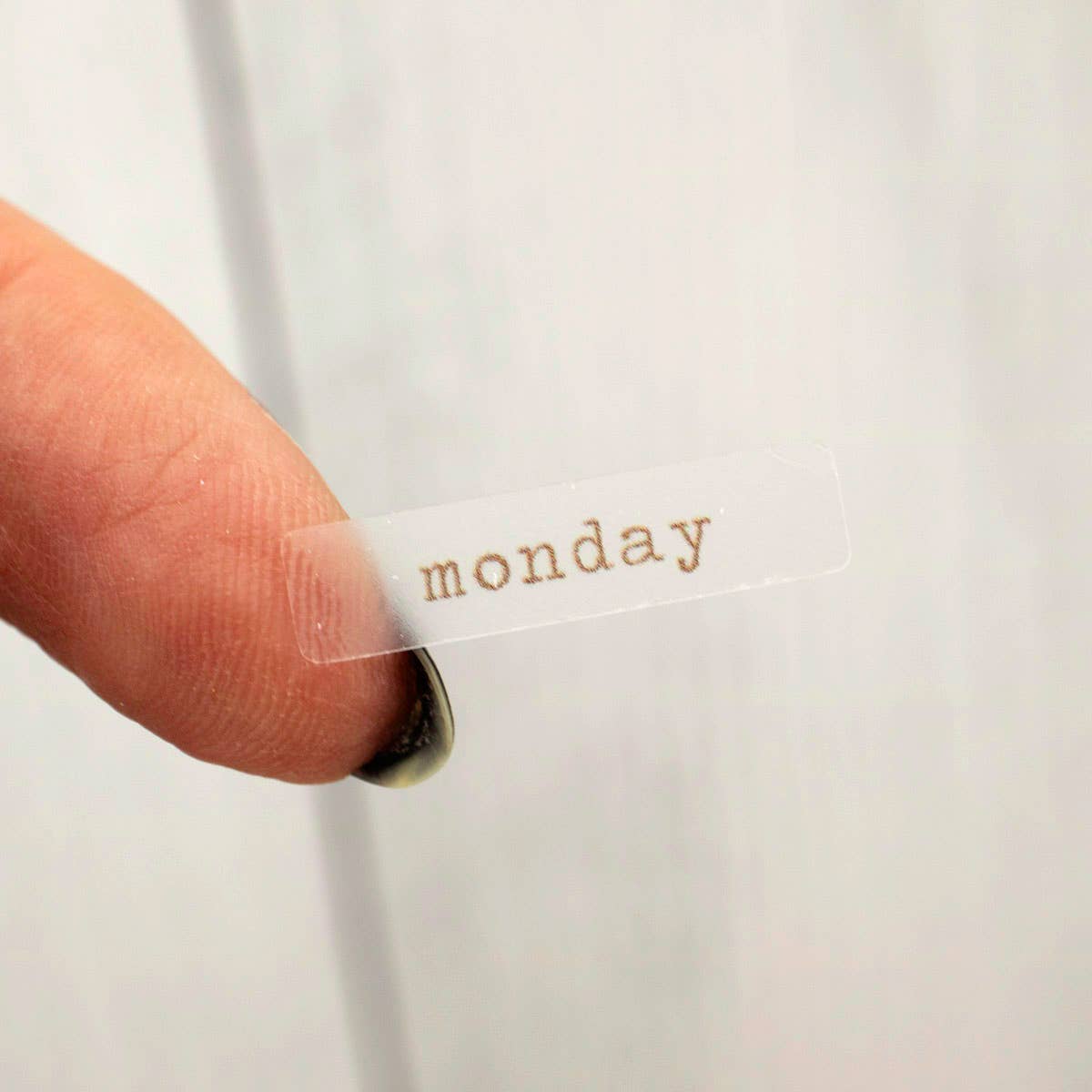 Clear adhesive strip with 'monday' text being pressed onto a white surface by a finger.