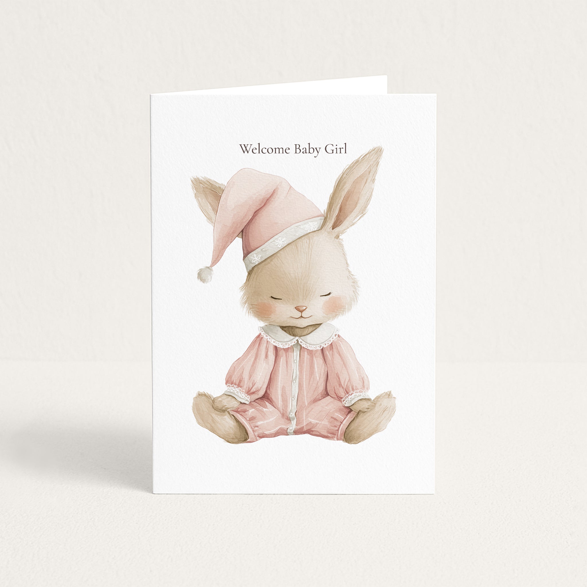 A white baby greeting card featuring a watercolor illustration of a sleeping bunny in pink pajamas and a matching hat, with the text 'Welcome Baby Girl' above it, shown on a light beige background.
