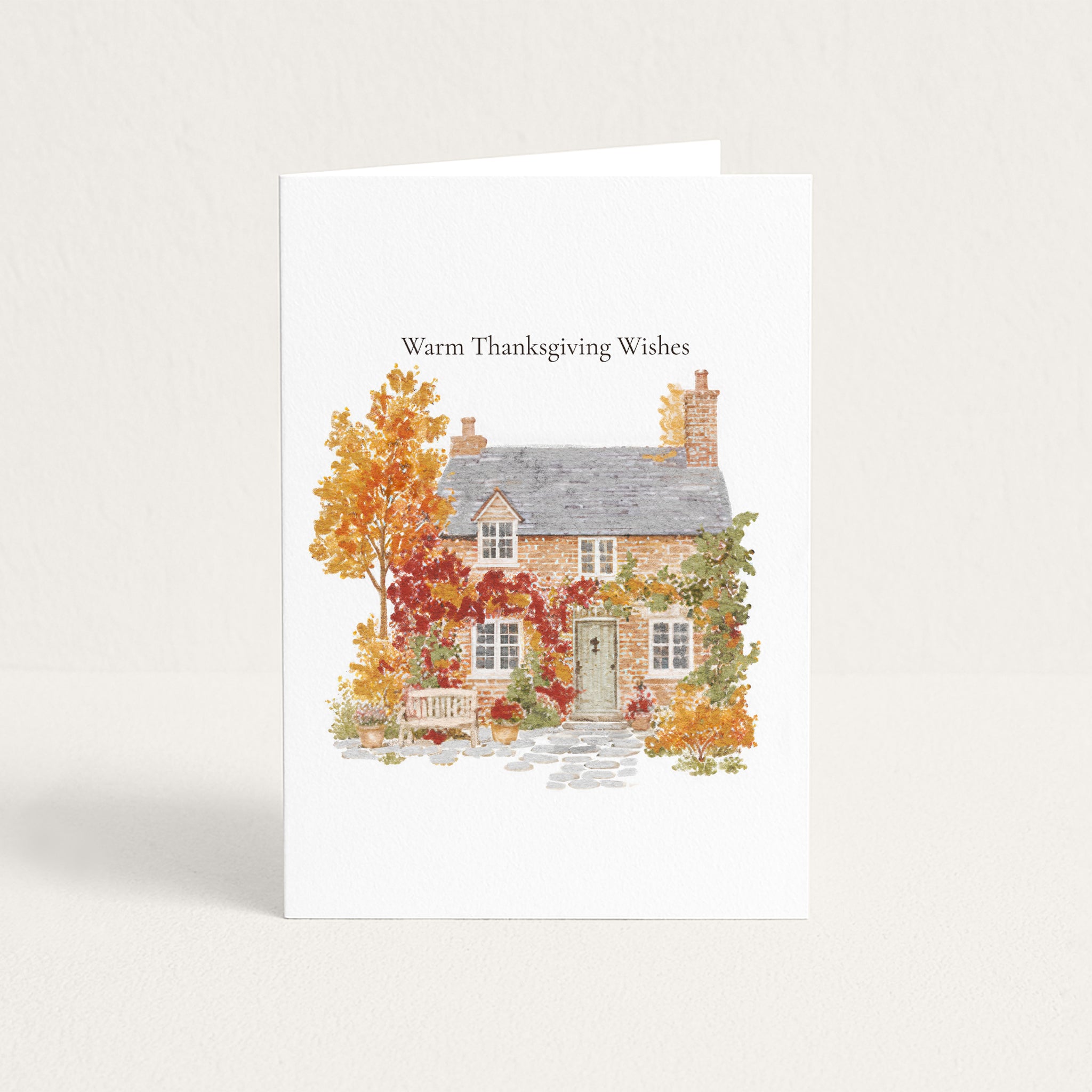 A white greeting card standing upright on a light neutral background. The card features the text 'Warm Thanksgiving Wishes' above a watercolor-style illustration of a small house surrounded by autumn foliage in orange, red, yellow, and green.
