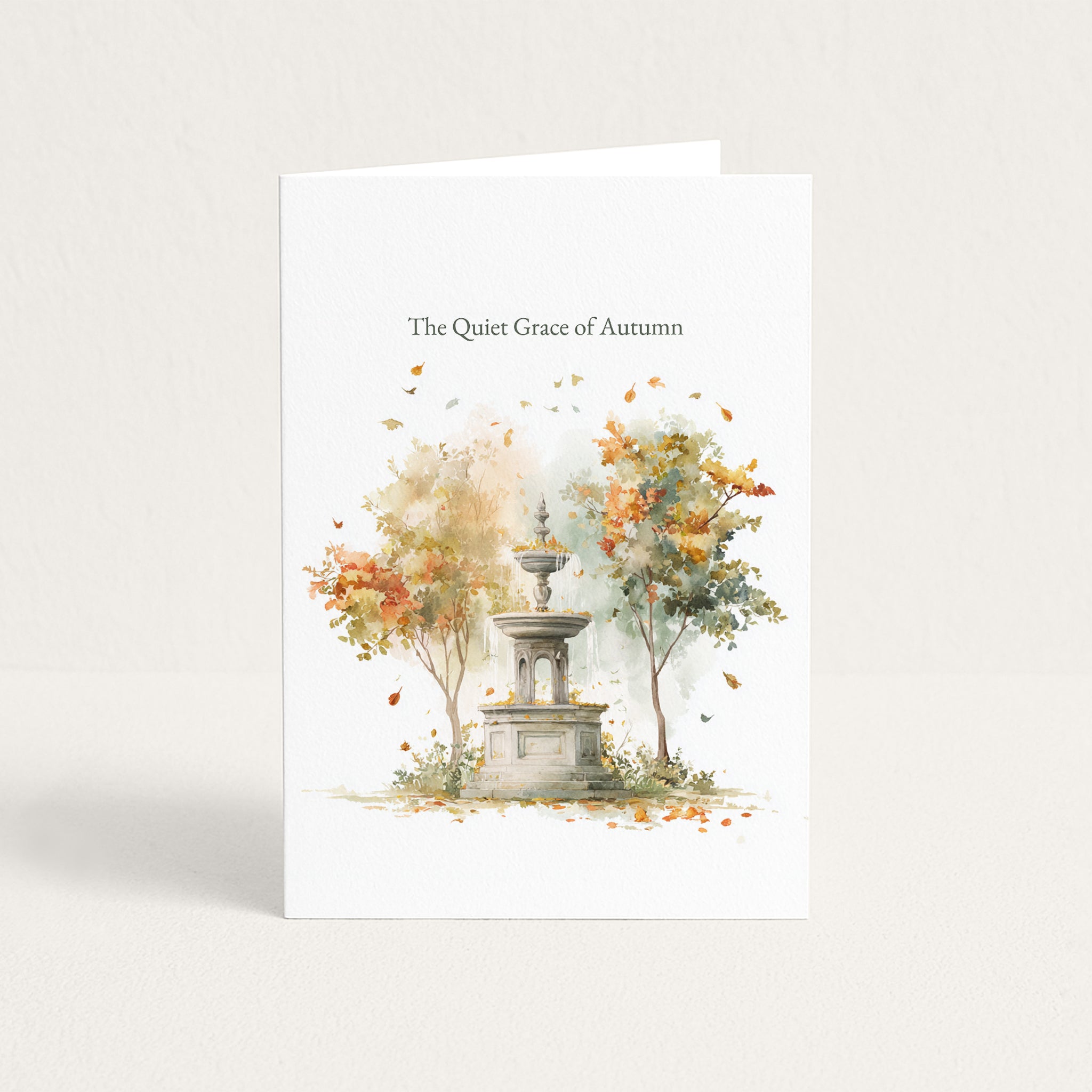 A white greeting card standing upright against a light neutral background. The card features a watercolor illustration of a stone fountain surrounded by autumn trees with falling leaves, and the text 'The Quiet Grace of Autumn' printed near the top.
