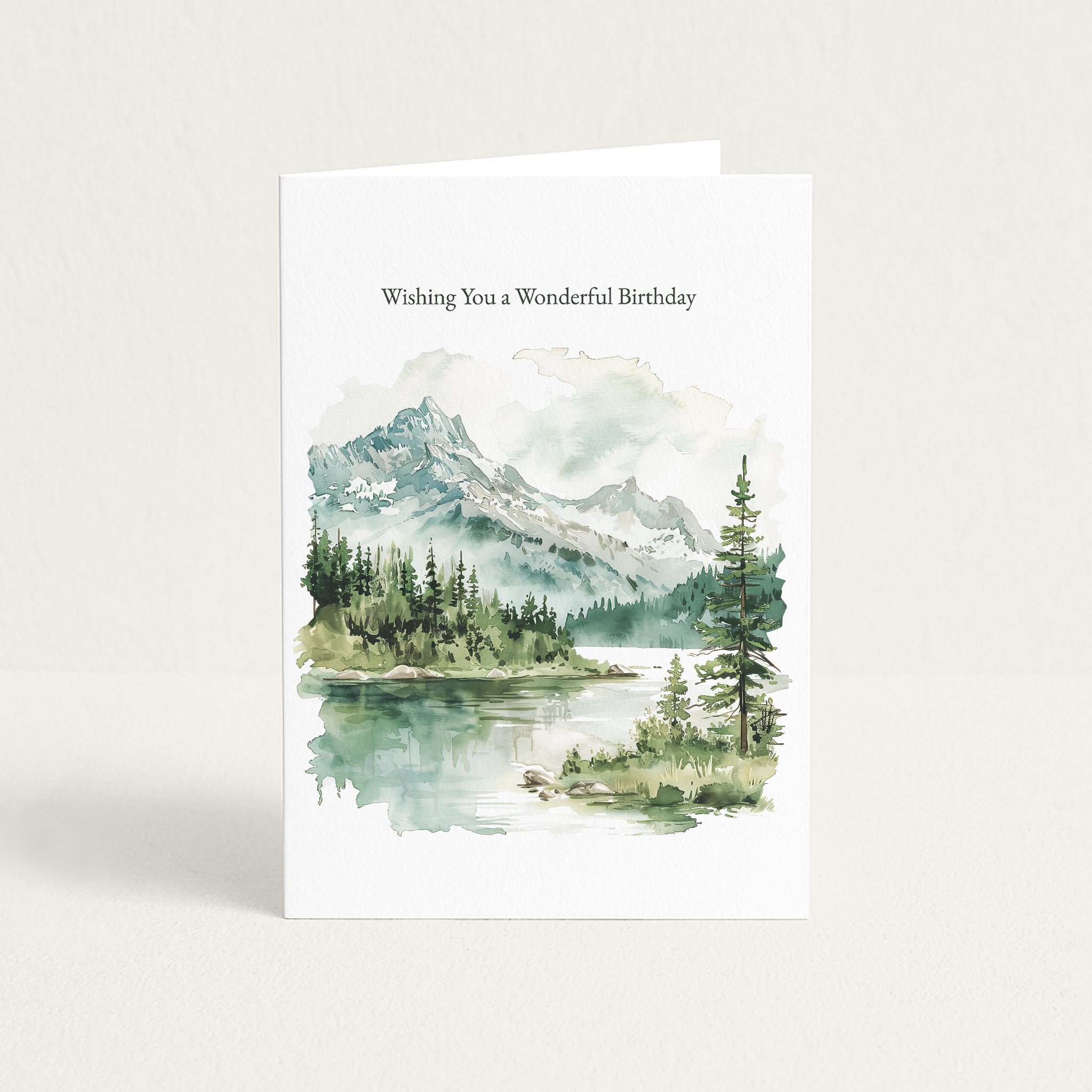 A white greeting card standing upright, printed with the text 'Wishing You a Wonderful Birthday' above a watercolor landscape of mountains, trees, and a reflective lake.