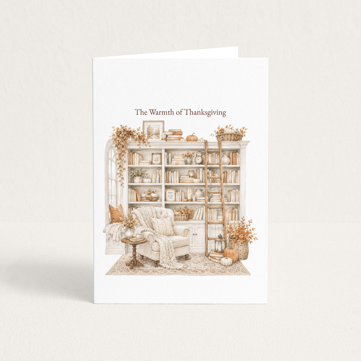 A white greeting card standing upright against a light beige background. The front of the card features an illustrated cozy library scene in muted warm tones, with the text 'The Warmth of Thanksgiving' above the artwork.