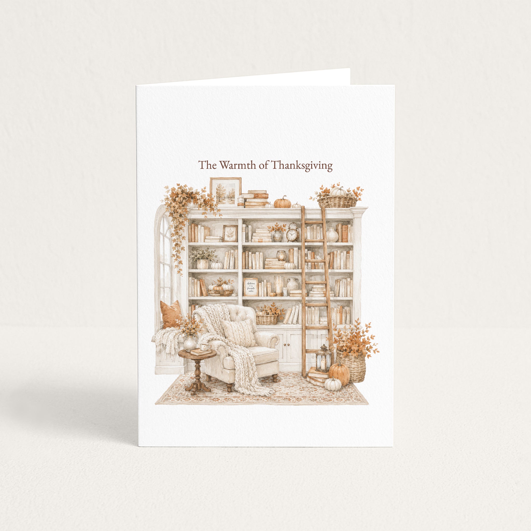 A white greeting card standing upright against a light beige background. The front of the card features an illustrated cozy library scene in muted warm tones, with the text 'The Warmth of Thanksgiving' above the artwork.