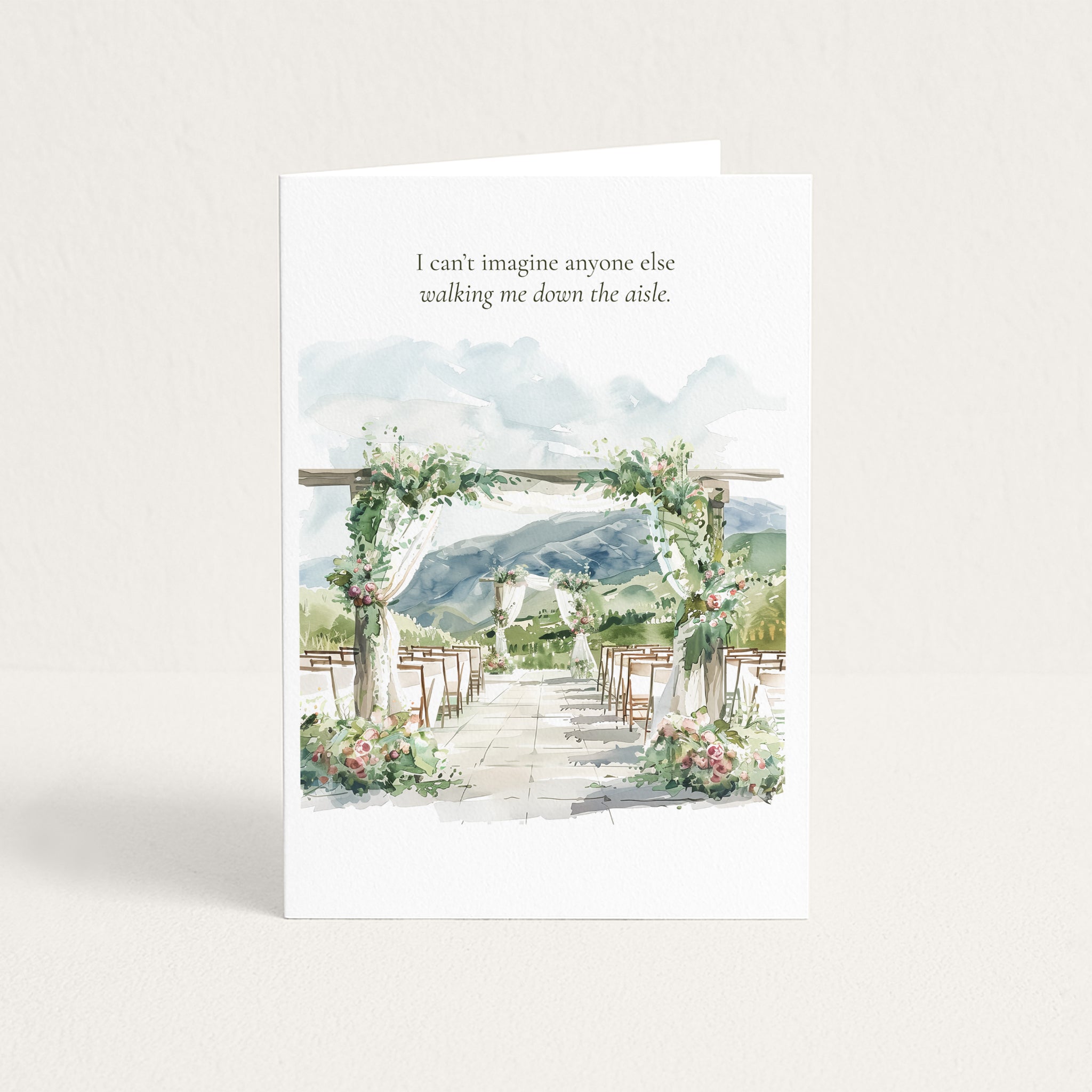 A white greeting card standing upright on a light beige surface. The card features a watercolor illustration of a wedding aisle with floral arches, chairs, and mountains in the background, with the printed text: 'I can't imagine anyone else walking me down the aisle.