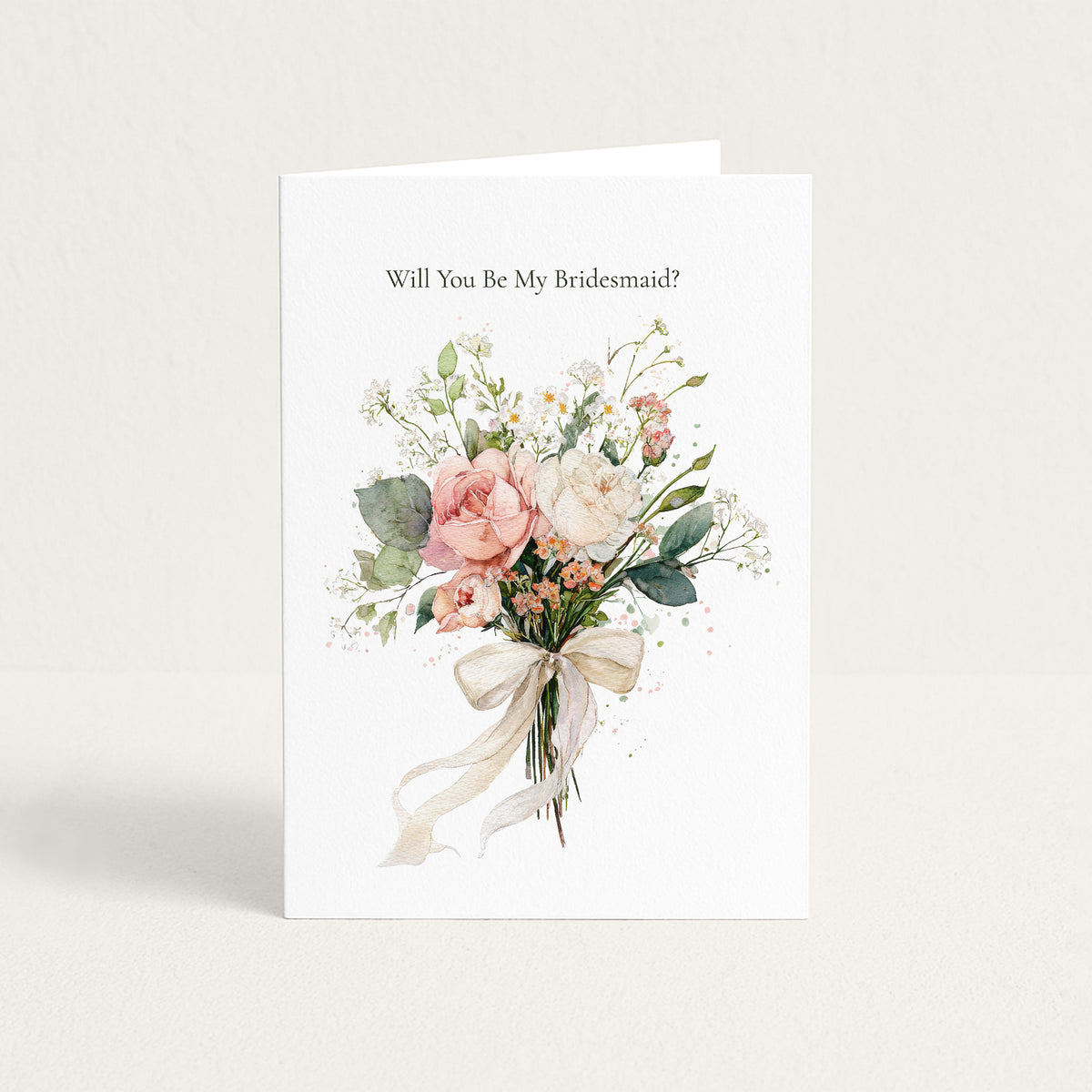 A white bridal shower or bachelorette greeting card standing upright, printed with the text 'Will You Be My Bridesmaid?' above a watercolor-style bouquet of blush pink, cream, and green flowers tied with a cream ribbon.