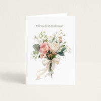 A white bridal shower or bachelorette greeting card standing upright, printed with the text 'Will You Be My Bridesmaid?' above a watercolor-style bouquet of blush pink, cream, and green flowers tied with a cream ribbon.