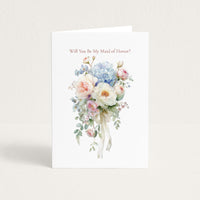 A white greeting card standing upright against a light beige background. The card features the text 'Will You Be My Maid of Honor?' above a delicate watercolor bouquet of pink, blue, and white flowers with green leaves and a white ribbon.