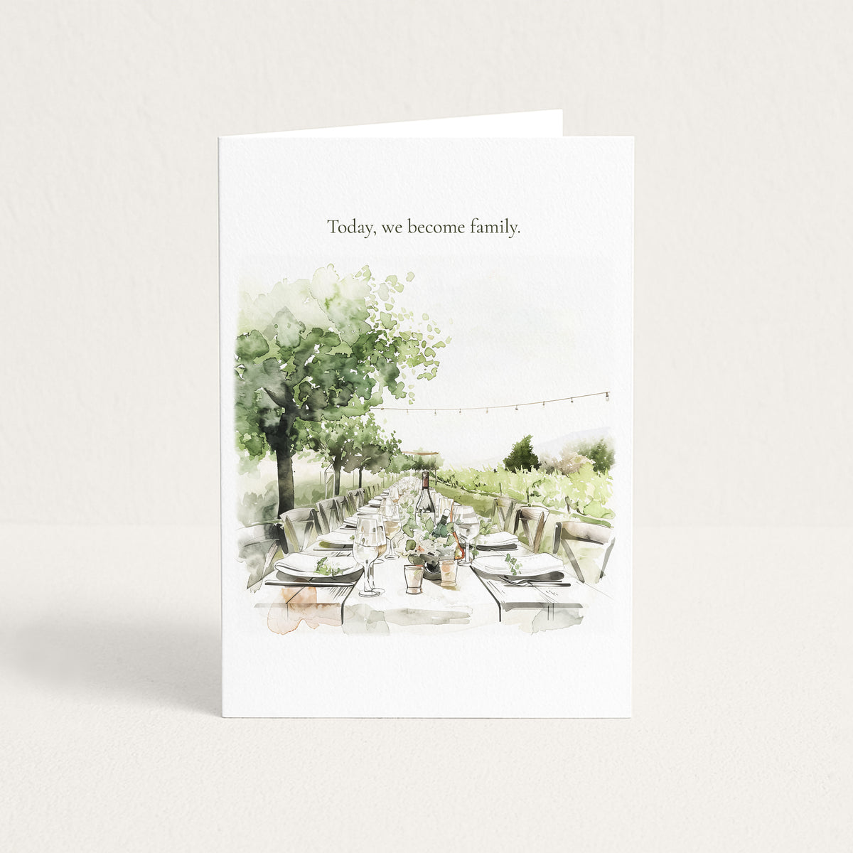 A white greeting card standing upright on a light beige background. The card features the printed text 'Today, we become family.