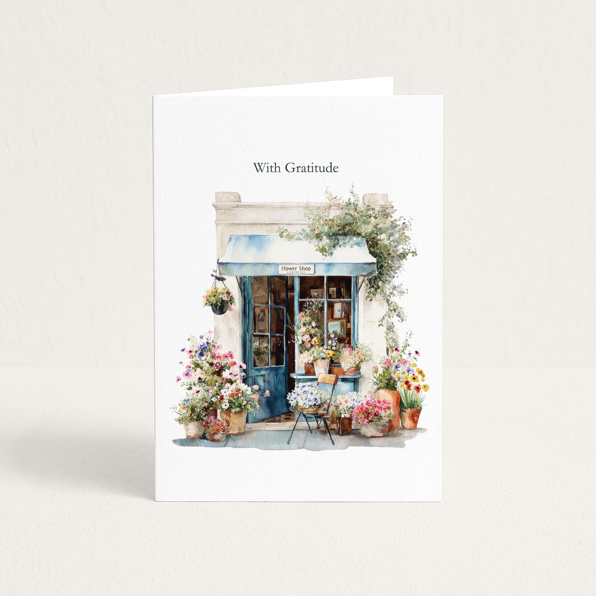 A white greeting card standing upright against a light beige background. The card features a watercolor illustration of a quaint flower shop with blue awnings, colorful flower arrangements, and the text 'With Gratitude' printed above the artwork.
