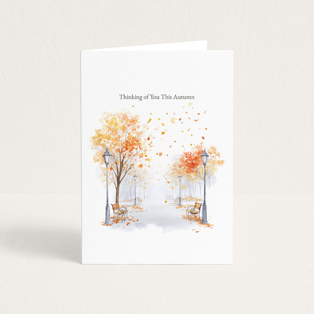 A white greeting card standing upright on a light beige surface, printed with the text 'Thinking of You This Autumn' above a watercolor scene of trees with orange and yellow leaves, benches, and street lamps in a misty park.