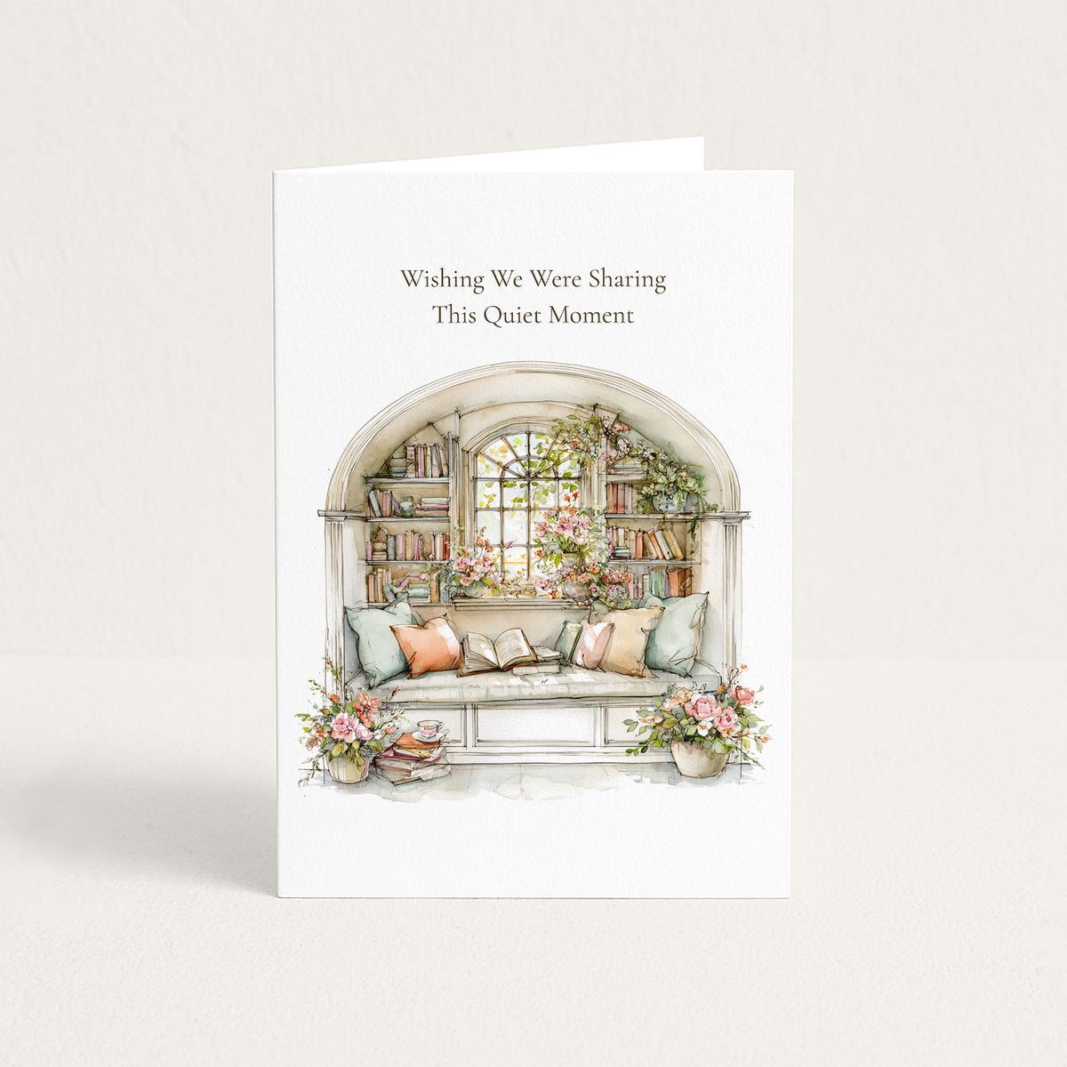 A white greeting card standing upright against a light neutral background. The card features the text 'Wishing We Were Sharing This Quiet Moment' above a watercolor-style illustration of a cozy window seat with pillows, books, flowers, and plants.