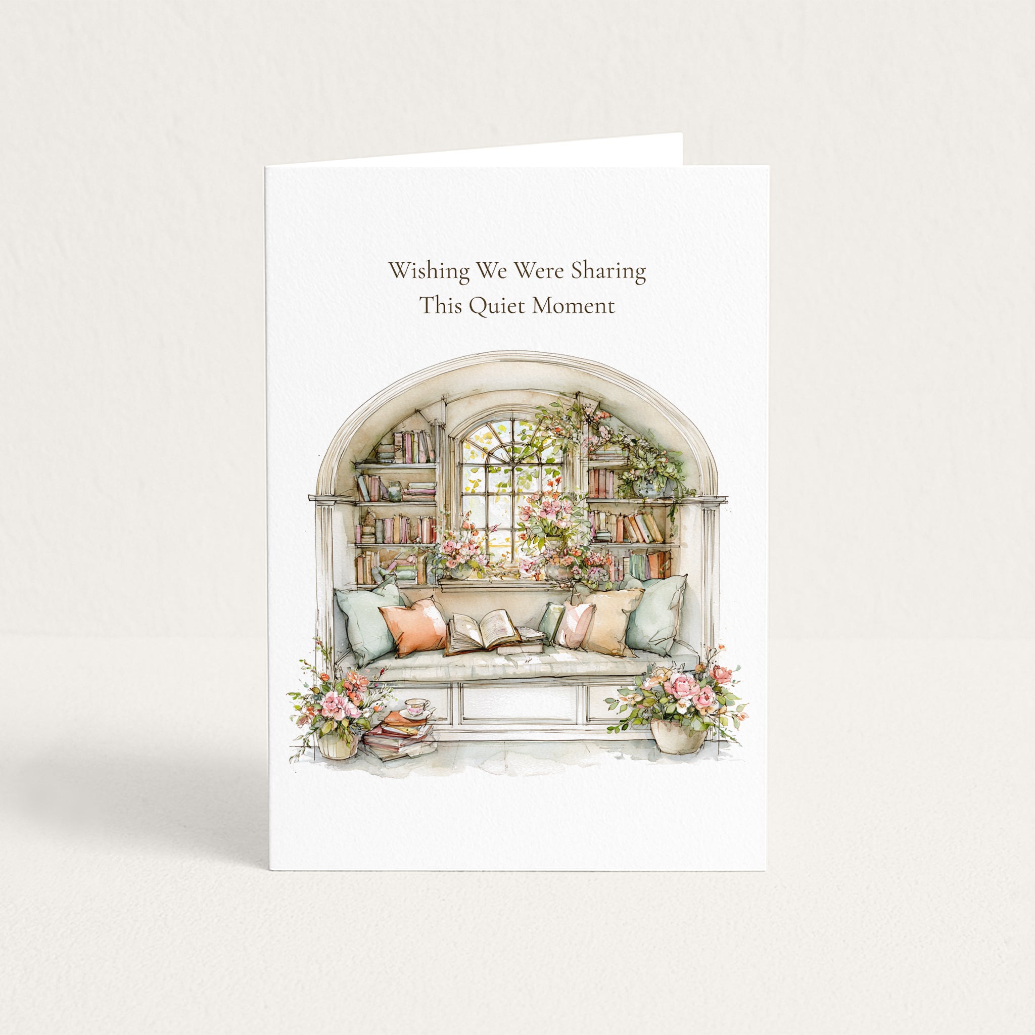 A white greeting card standing upright against a light neutral background. The card features the text 'Wishing We Were Sharing This Quiet Moment' above a watercolor-style illustration of a cozy window seat with pillows, books, flowers, and plants.