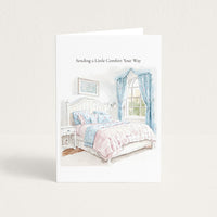A white greeting card standing upright on a light beige background. The card features a watercolor illustration of a cozy bed with pink and blue bedding, blue curtains, and the printed text 'Sending a Little Comfort Your Way' at the top.