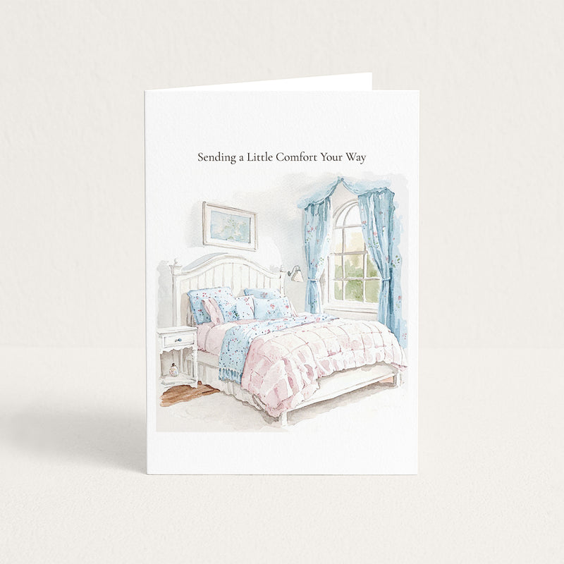 A white greeting card standing upright on a light beige background. The card features a watercolor illustration of a cozy bed with pink and blue bedding, blue curtains, and the printed text 'Sending a Little Comfort Your Way' at the top.