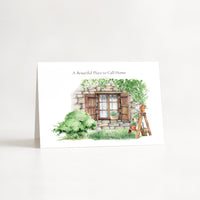 A vertical white greeting card standing upright on a white surface against a white background. The card features a watercolor illustration of a rustic stone cottage with green bushes, a wooden window, and a small garden cart, with the text 'A Beautiful Place to Call Home' printed above the artwork.