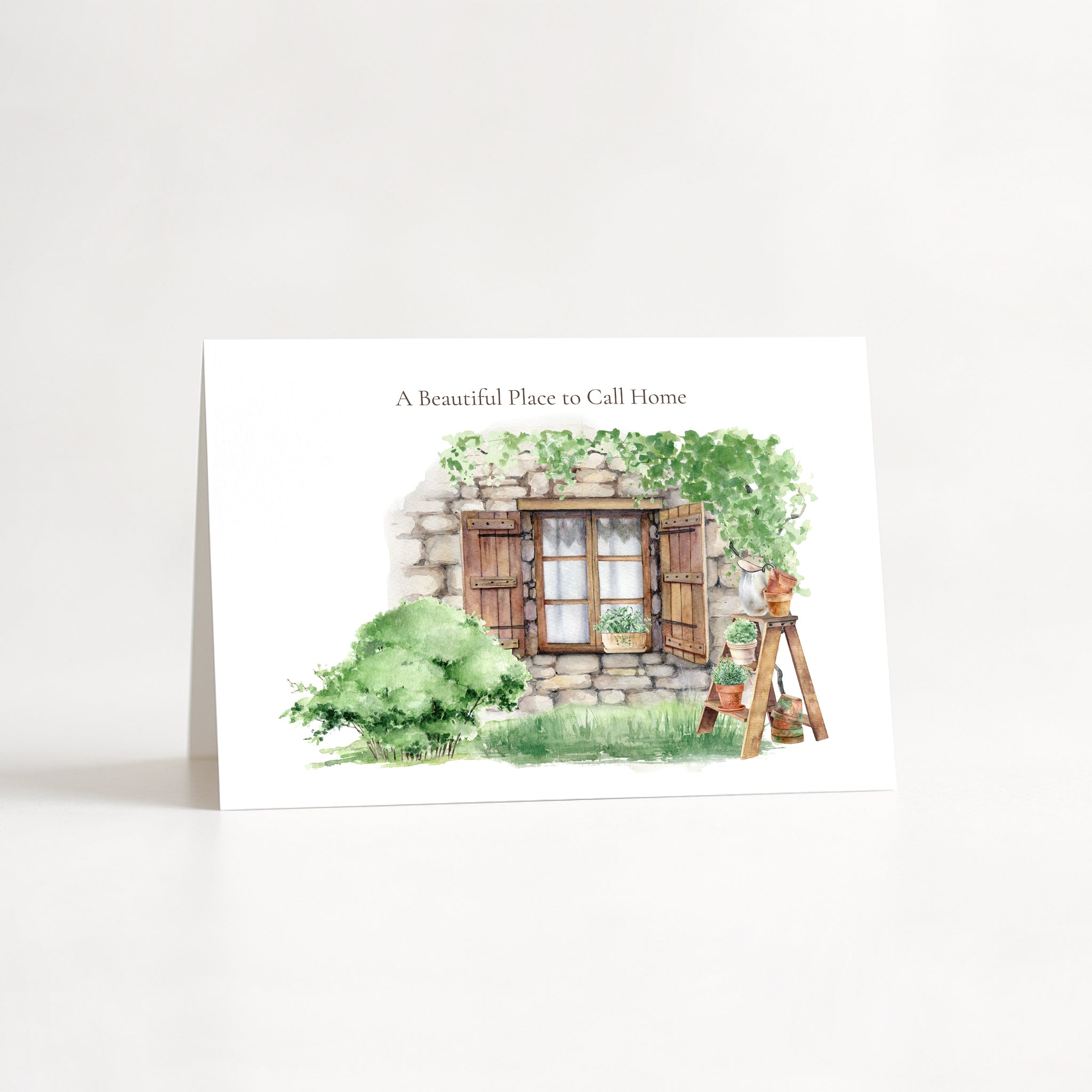 A vertical white greeting card standing upright on a white surface against a white background. The card features a watercolor illustration of a rustic stone cottage with green bushes, a wooden window, and a small garden cart, with the text 'A Beautiful Place to Call Home' printed above the artwork.