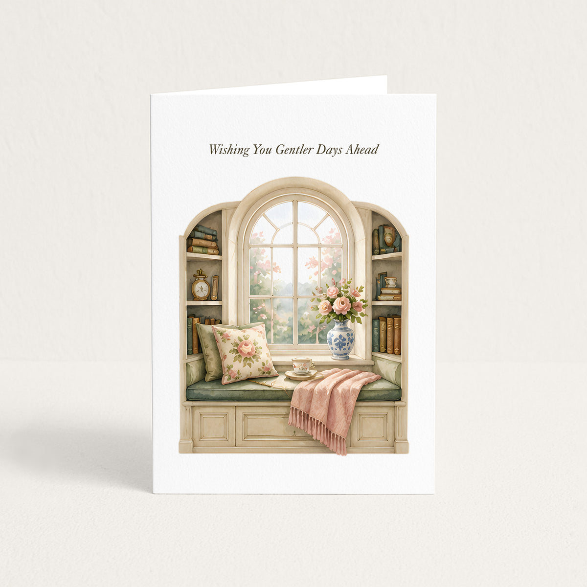 A white greeting card standing upright against a light beige background. The card features an illustration of a cozy window seat with floral pillows, a pink throw blanket, books on the shelves, and a vase of flowers, with the text 'Wishing You Gentler Days Ahead' printed above the illustration.