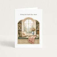A white greeting card standing upright against a light beige background. The card features an illustration of a cozy window seat with floral pillows, a pink throw blanket, books on the shelves, and a vase of flowers, with the text 'Wishing You Gentler Days Ahead' printed above the illustration.