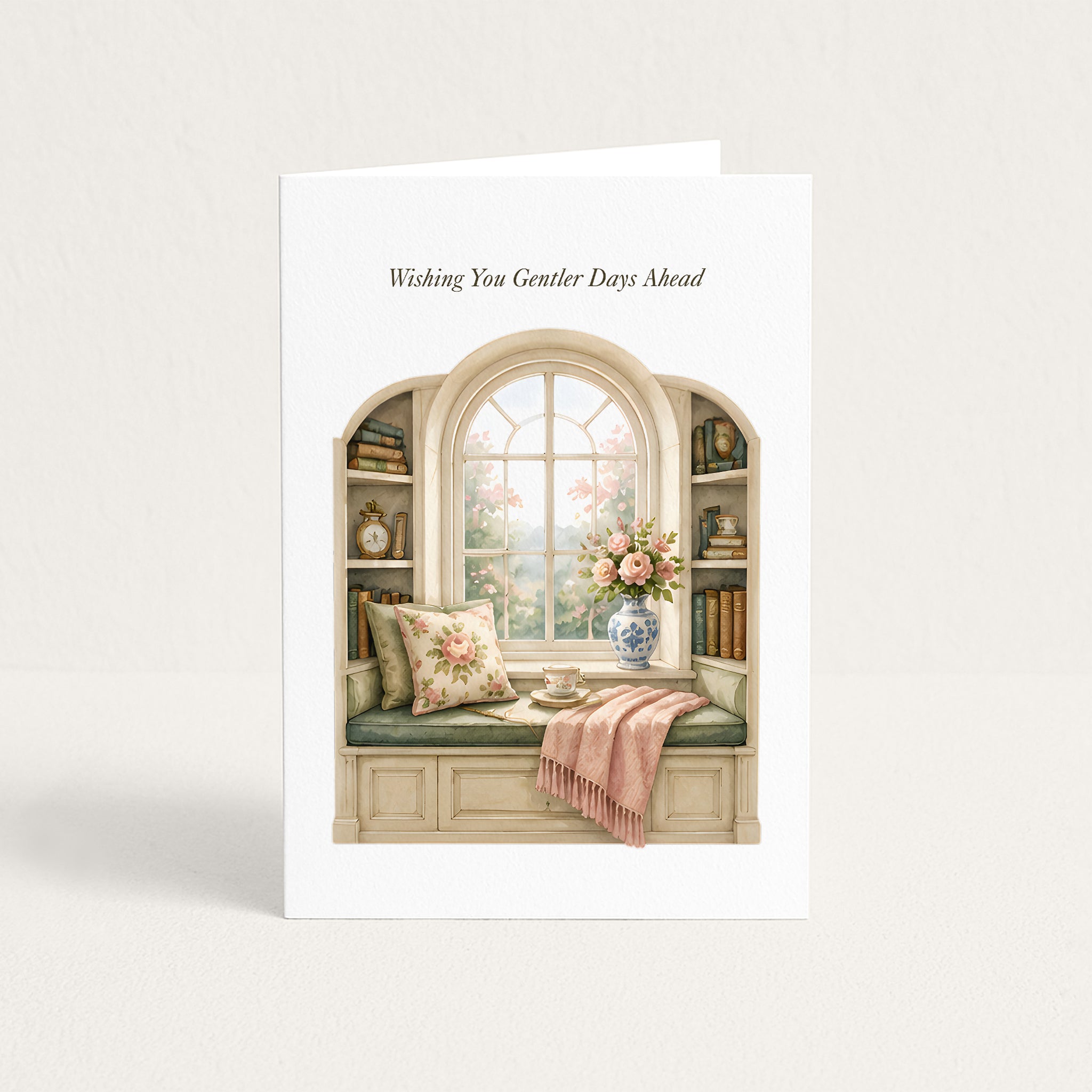 A white greeting card standing upright against a light beige background. The card features an illustration of a cozy window seat with floral pillows, a pink throw blanket, books on the shelves, and a vase of flowers, with the text 'Wishing You Gentler Days Ahead' printed above the illustration.
