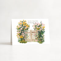 A white greeting card standing upright on a white surface, featuring a watercolor-style illustration of a small white picket fence surrounded by pink, yellow, and green flowers. Small black text at the top reads, 'You Have Always Made Life Beautiful.