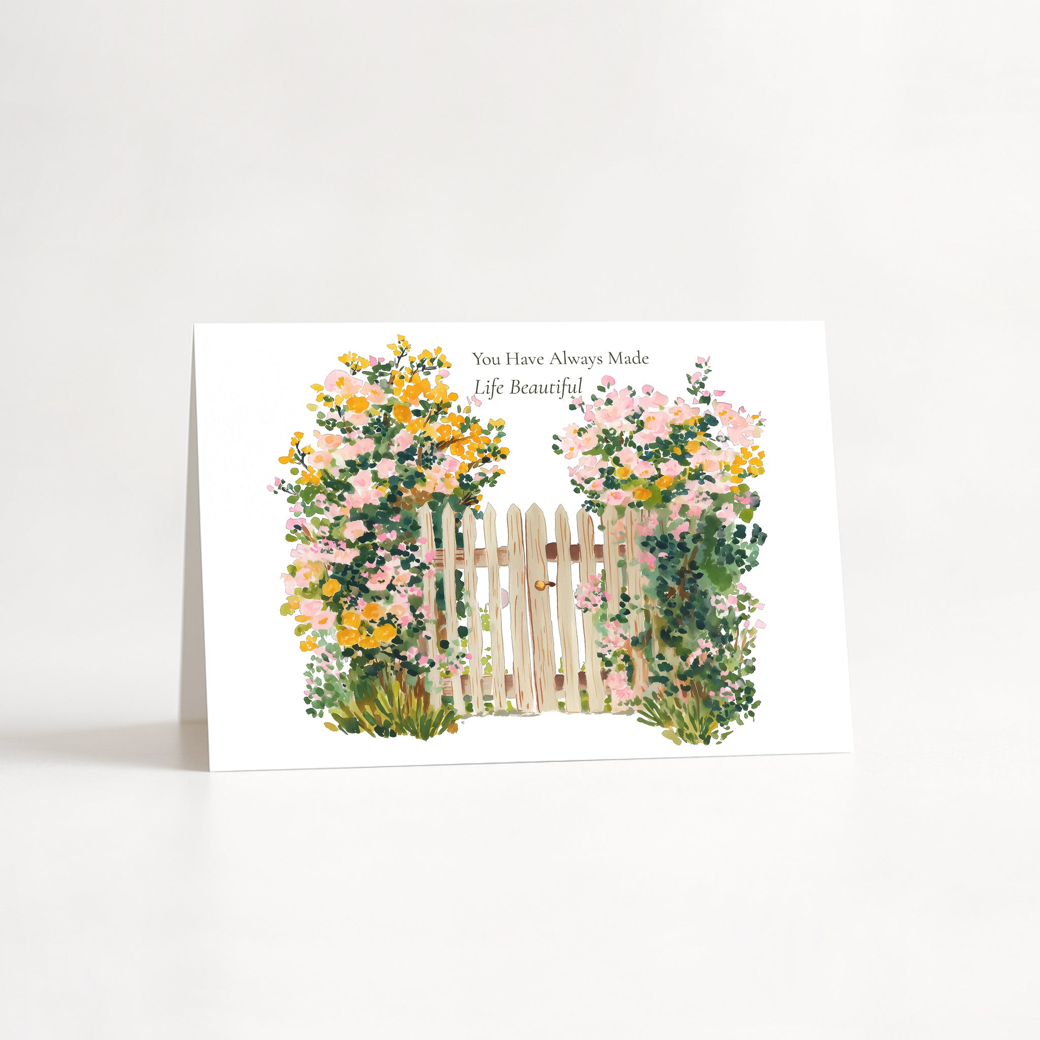 A white greeting card standing upright on a white surface, featuring a watercolor-style illustration of a small white picket fence surrounded by pink, yellow, and green flowers. Small black text at the top reads, 'You Have Always Made Life Beautiful.