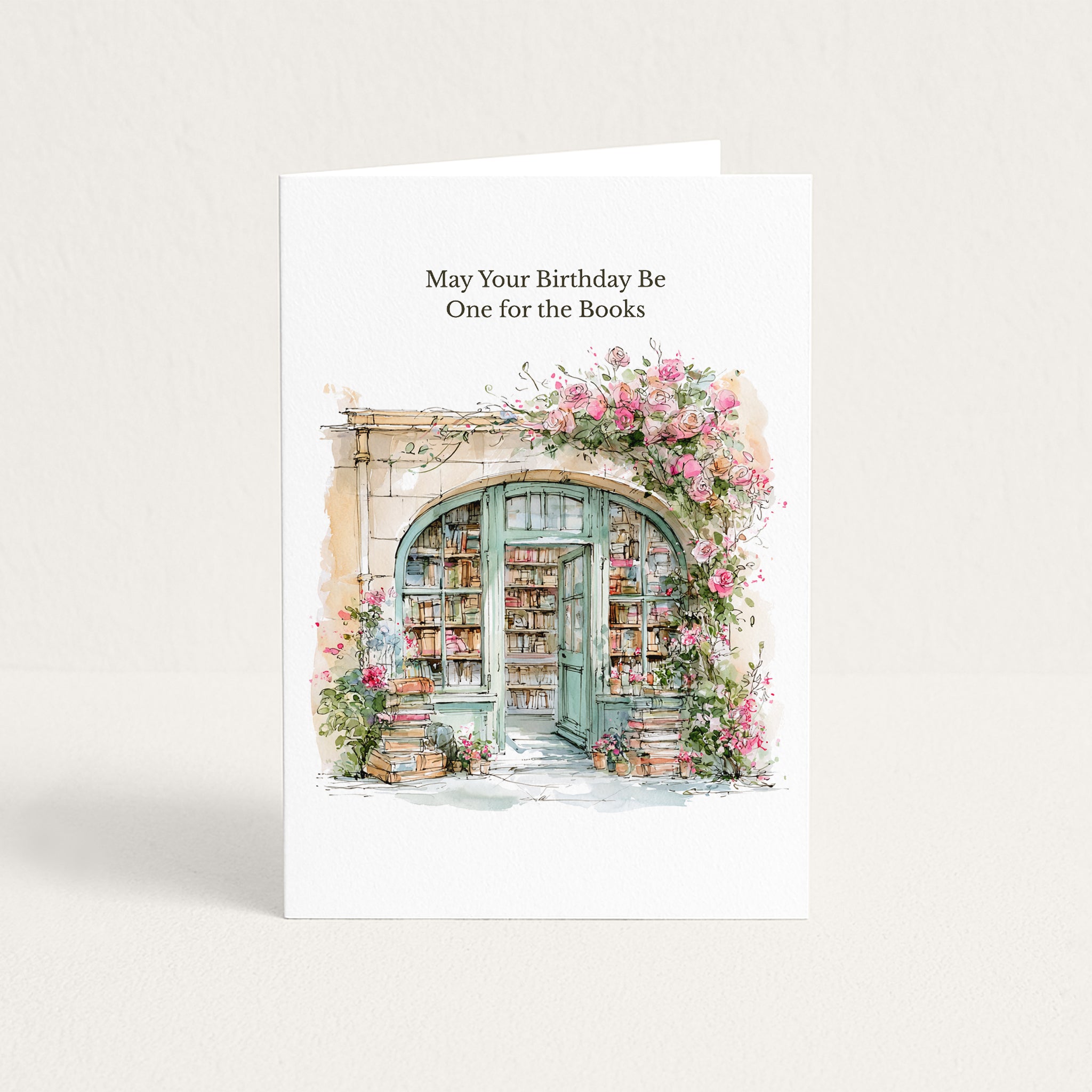 A white greeting card standing upright on a light neutral background. The card features the printed text 'May Your Birthday Be One for the Books' above a watercolor-style illustration of an open bookshop door framed by pink flowers and books.
