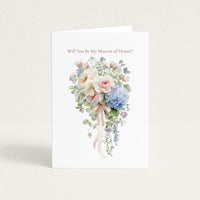 A white greeting card standing upright on a light beige surface, printed with the phrase 'Will You Be My Marron of Honor?' above a watercolor-style bouquet in soft pink, cream, blue, and green tones.