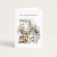 A white greeting card standing upright against a light beige background. The card features the printed text 'How Lovely to Hear From You' above a watercolor-style illustration of a window, flowers, a chair, and stacked books.
