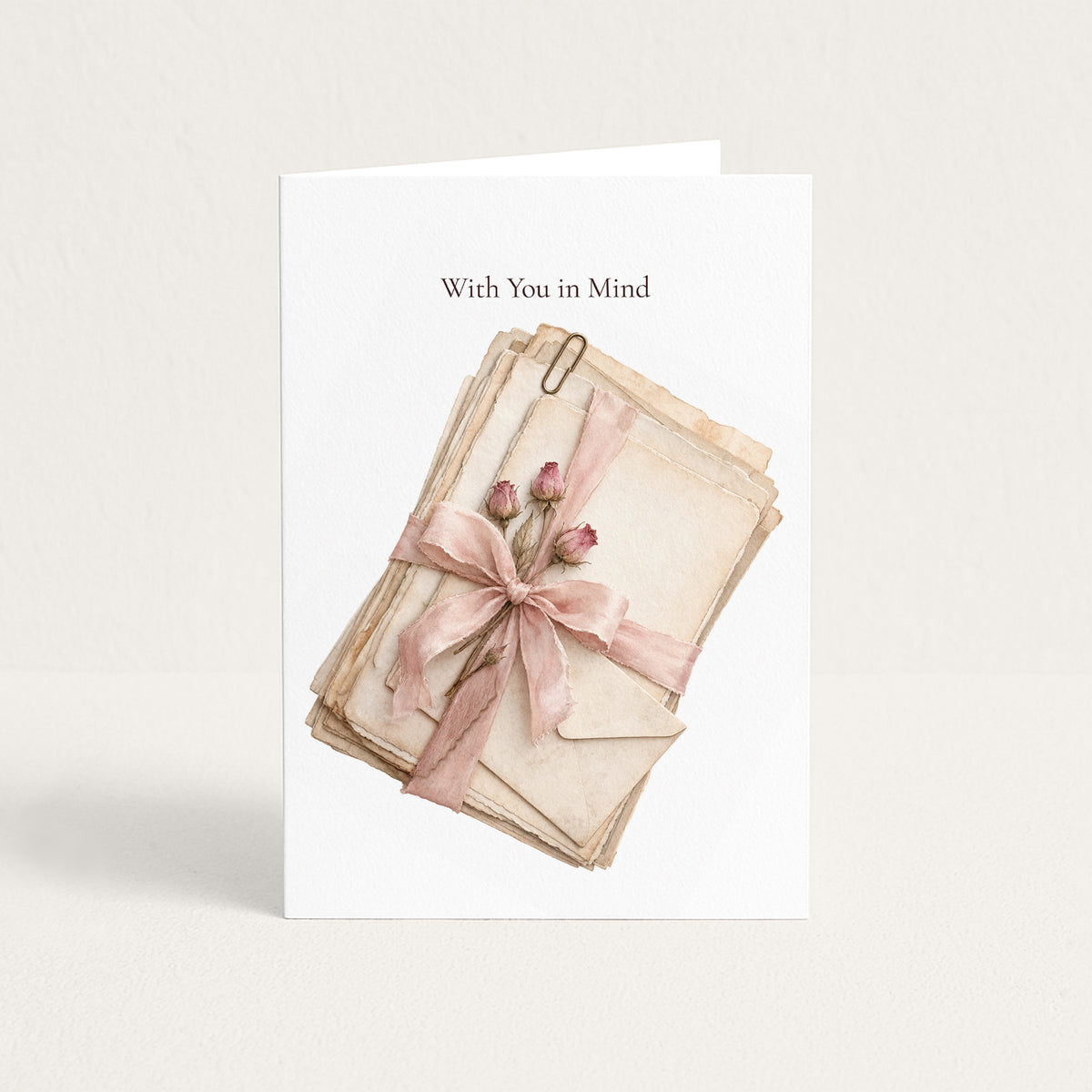 A white greeting card standing upright on a light beige background. The card has the words 'With You in Mind' printed at the top and features an illustration of stacked vintage envelopes tied with a pink ribbon and bow, with small pink flower buds.