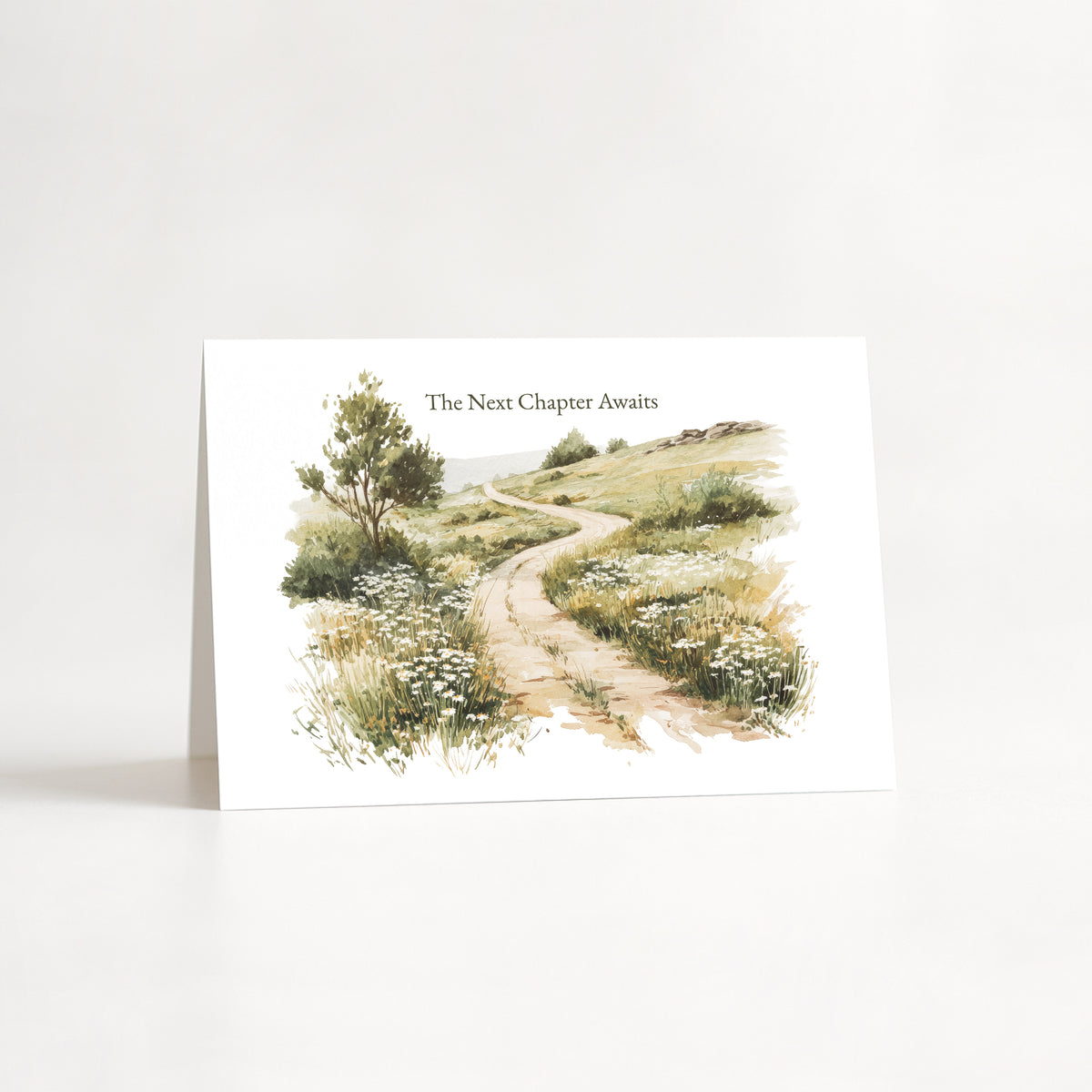 A white greeting card standing upright on a white surface, featuring a watercolor illustration of a winding dirt path through grassy hills with a tree on the left. The text 'The Next Chapter Awaits' appears above the artwork.