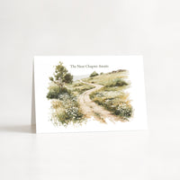 A white greeting card standing upright on a white surface, featuring a watercolor illustration of a winding dirt path through grassy hills with a tree on the left. The text 'The Next Chapter Awaits' appears above the artwork.