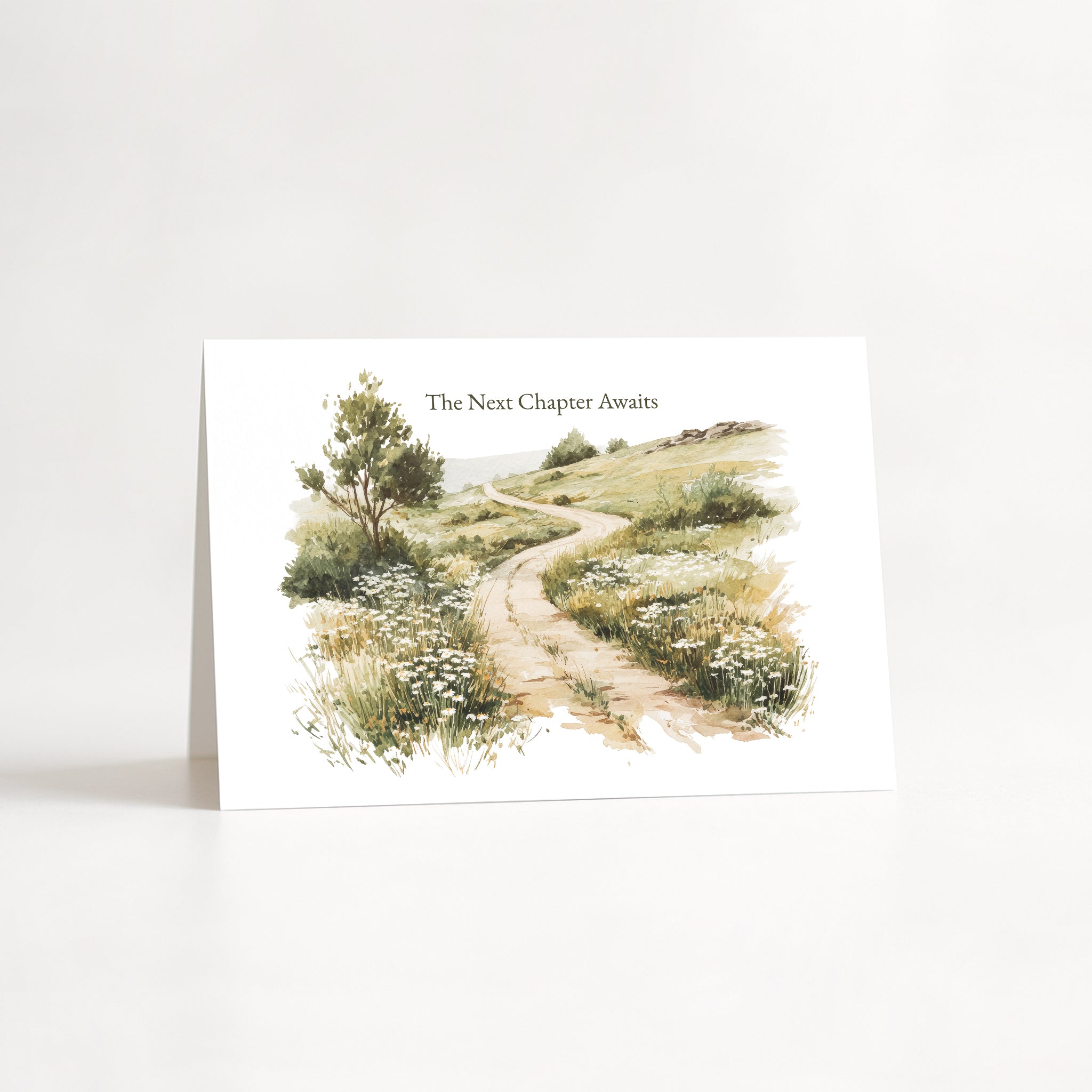 A white greeting card standing upright on a white surface, featuring a watercolor illustration of a winding dirt path through grassy hills with a tree on the left. The text 'The Next Chapter Awaits' appears above the artwork.