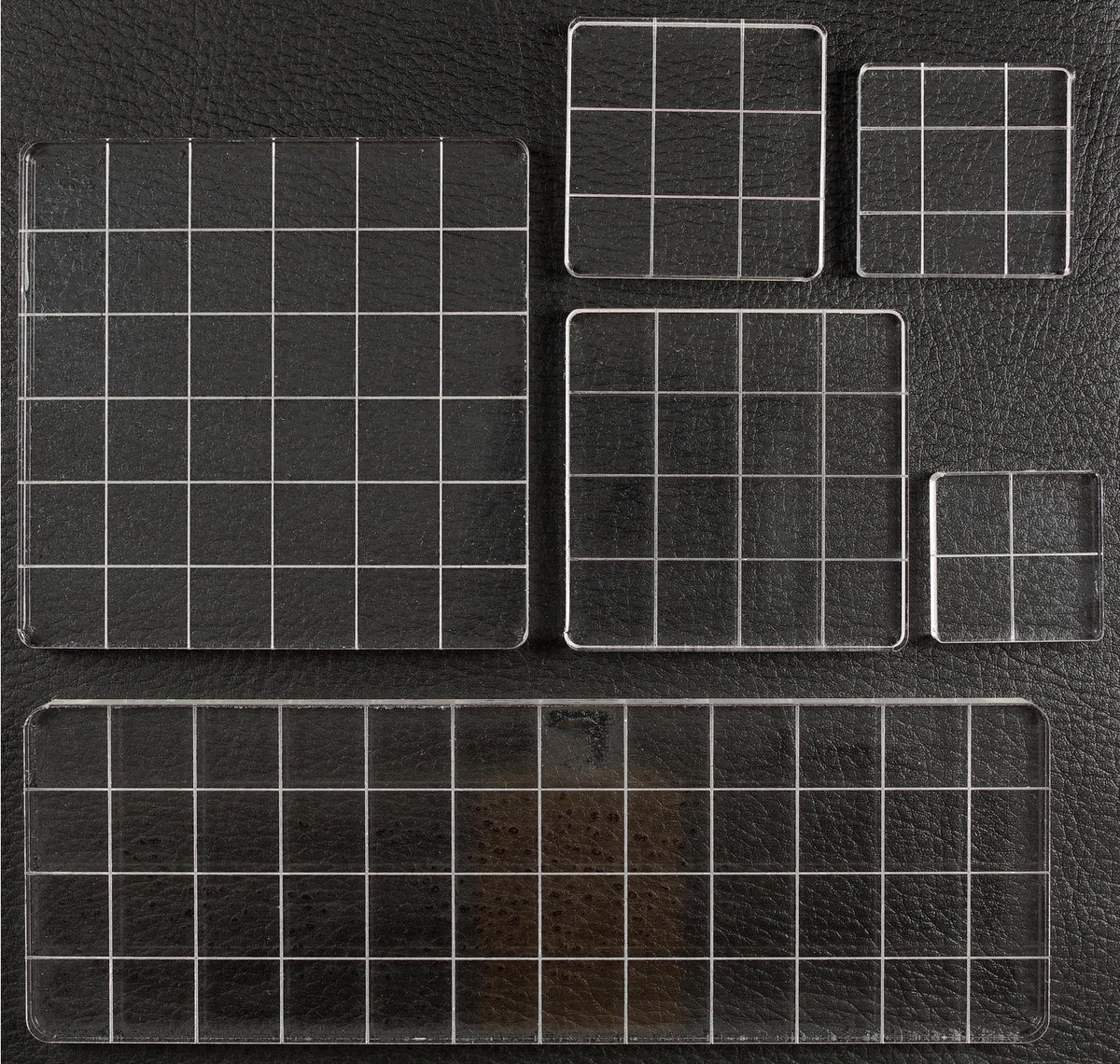 A set of clear acrylic or glass grid trays with white grid lines, shown in multiple sizes on a black textured background.