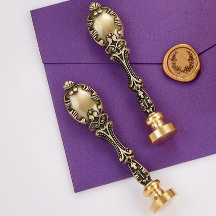 Two ornate antique-style wax seal stamps with decorative handles and gold-colored seals, displayed on a purple envelope against a white background.