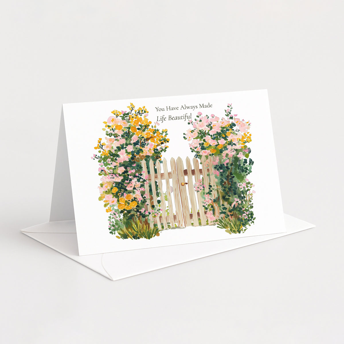 A white greeting card standing open on a white envelope against a light gray background. The card features a watercolor-style illustration of a small wooden fence flanked by pink and yellow flowers, with the printed text 'You Have Always Made Life Beautiful'.