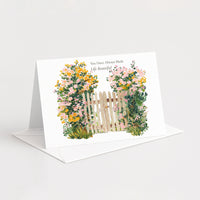 A white greeting card standing open on a white envelope against a light gray background. The card features a watercolor-style illustration of a small wooden fence flanked by pink and yellow flowers, with the printed text 'You Have Always Made Life Beautiful'.