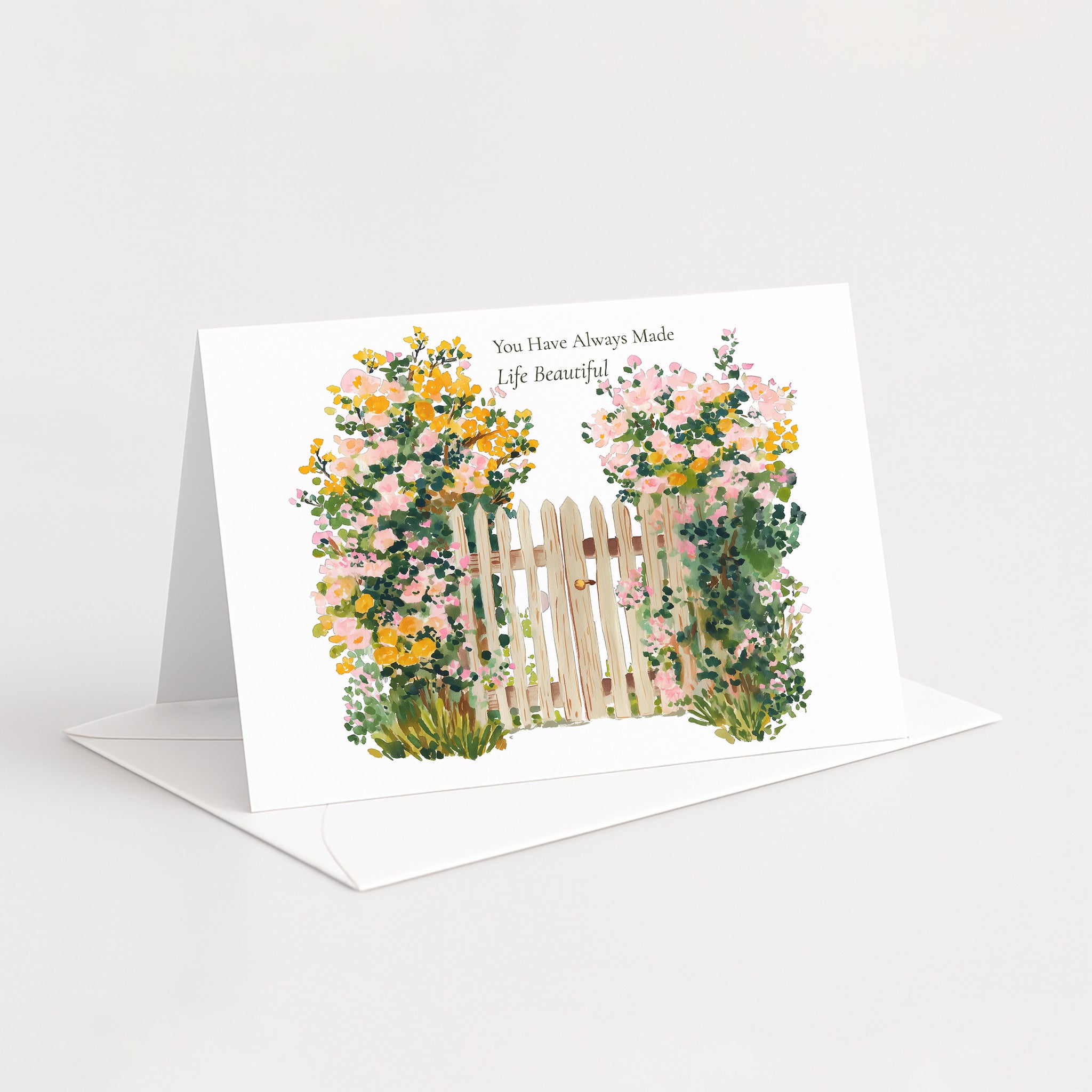 A white greeting card standing open on a white envelope against a light gray background. The card features a watercolor-style illustration of a small wooden fence flanked by pink and yellow flowers, with the printed text 'You Have Always Made Life Beautiful'.