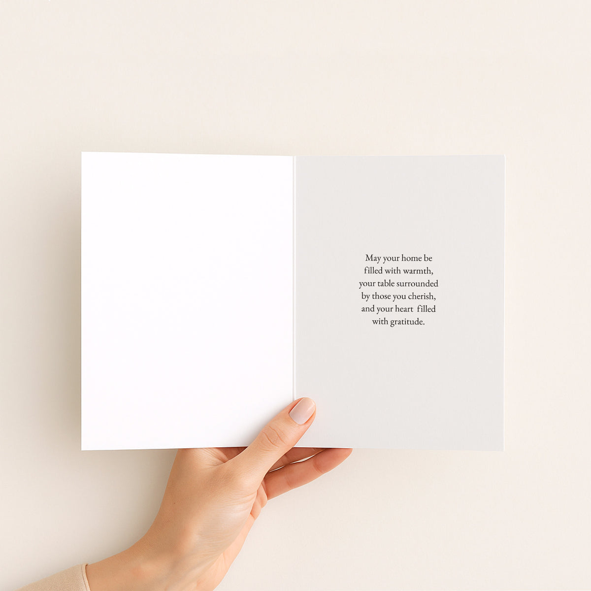 A hand holds an open white greeting card against a warm off-white background. The right page features a short, elegant black serif quote.