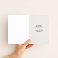 A hand holds an open white greeting card against a warm off-white background. The right page features a short, elegant black serif quote.