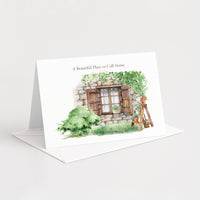 A white standing greeting card with an illustrated watercolor scene of a stone house window, greenery, and a ladder, with the text 'A Beautiful Place to Call Home.' It is shown standing upright with an envelope behind it on a clean white background.