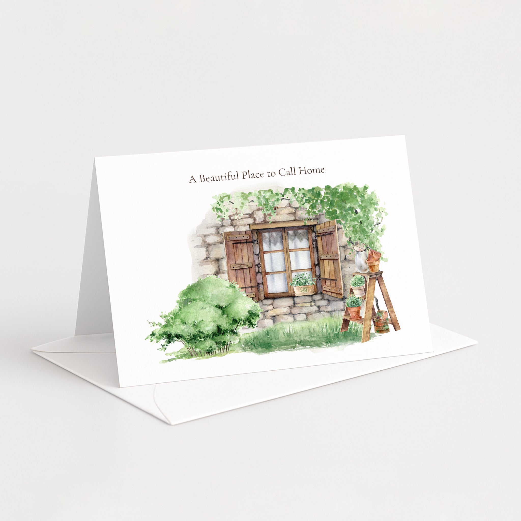 A white standing greeting card with an illustrated watercolor scene of a stone house window, greenery, and a ladder, with the text 'A Beautiful Place to Call Home.' It is shown standing upright with an envelope behind it on a clean white background.