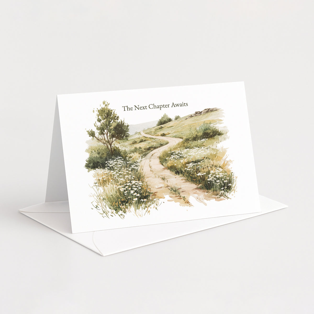 A white standing greeting card with an illustrated landscape scene of a winding path through grassy hills, shown open with a white envelope on a light gray background. The card reads 'The Next Chapter Awaits'.