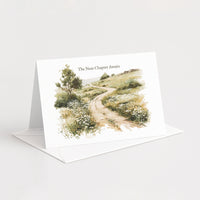 A white standing greeting card with an illustrated landscape scene of a winding path through grassy hills, shown open with a white envelope on a light gray background. The card reads 'The Next Chapter Awaits'.
