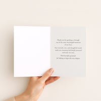An open white greeting card held by a hand against a warm off-white background. The right page contains a printed message in dark gray text, and the left page is blank.