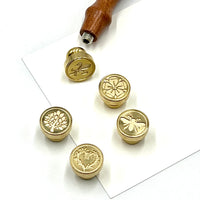 Five round brass stamp seals with engraved floral and insect designs are arranged on a white sheet, with a wooden-handled stamp tool at the top.