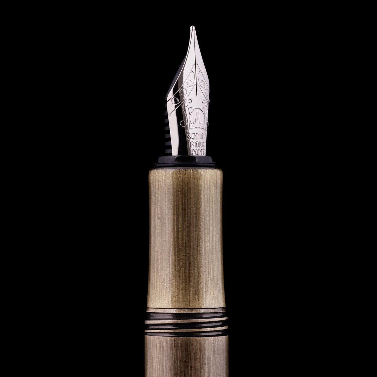 Close-up of a fountain pen nib and upper barrel against a solid black background. The nib is silver-toned with engraved lettering, and the barrel has a brushed gold/brass finish with black accent bands.