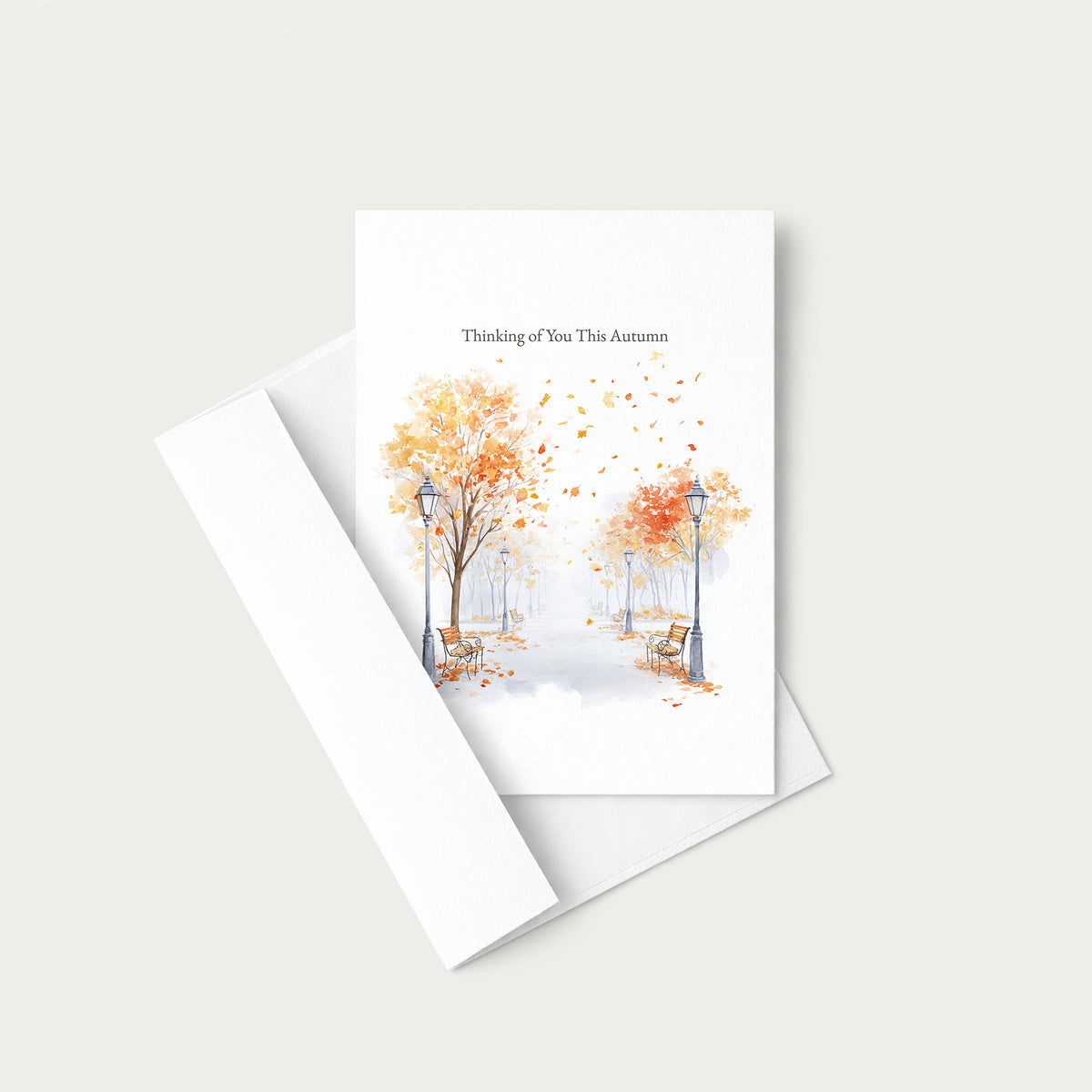 A white greeting card with a watercolor autumn scene of trees with orange and yellow leaves, benches, and street lamps, with the text 'Thinking of You This Autumn' at the top. The card is shown with its matching white envelope on a light beige background.