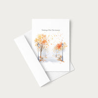 A white greeting card with a watercolor autumn scene of trees with orange and yellow leaves, benches, and street lamps, with the text 'Thinking of You This Autumn' at the top. The card is shown with its matching white envelope on a light beige background.