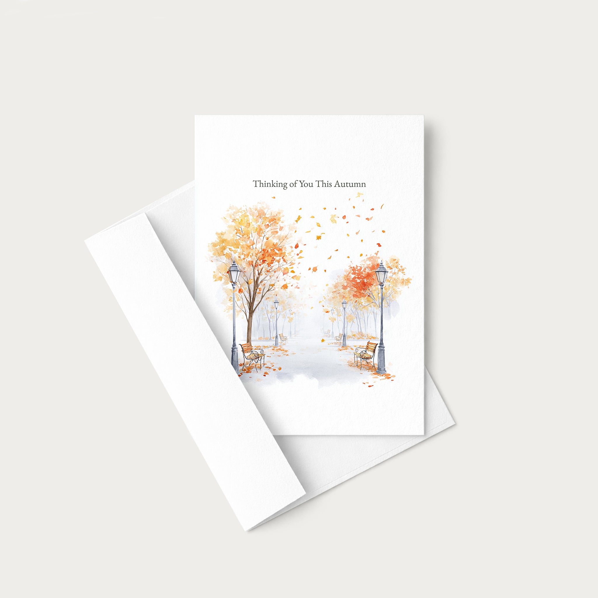 A white greeting card with a watercolor autumn scene of trees with orange and yellow leaves, benches, and street lamps, with the text 'Thinking of You This Autumn' at the top. The card is shown with its matching white envelope on a light beige background.